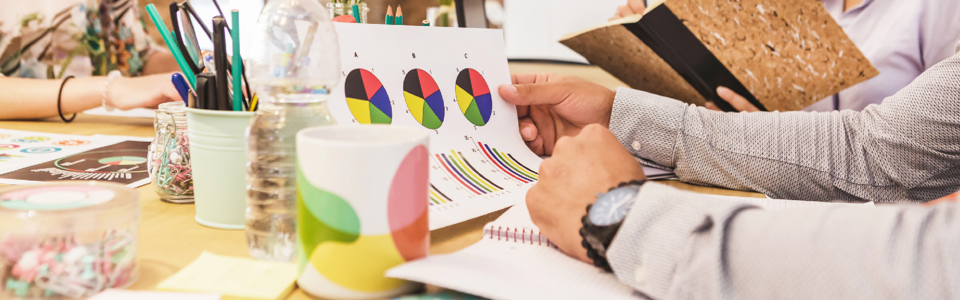 Team of professionals reviewing colorful pie charts and bar graphs during a meeting, surrounded by stationery and office supplies on a collaborative workspace.